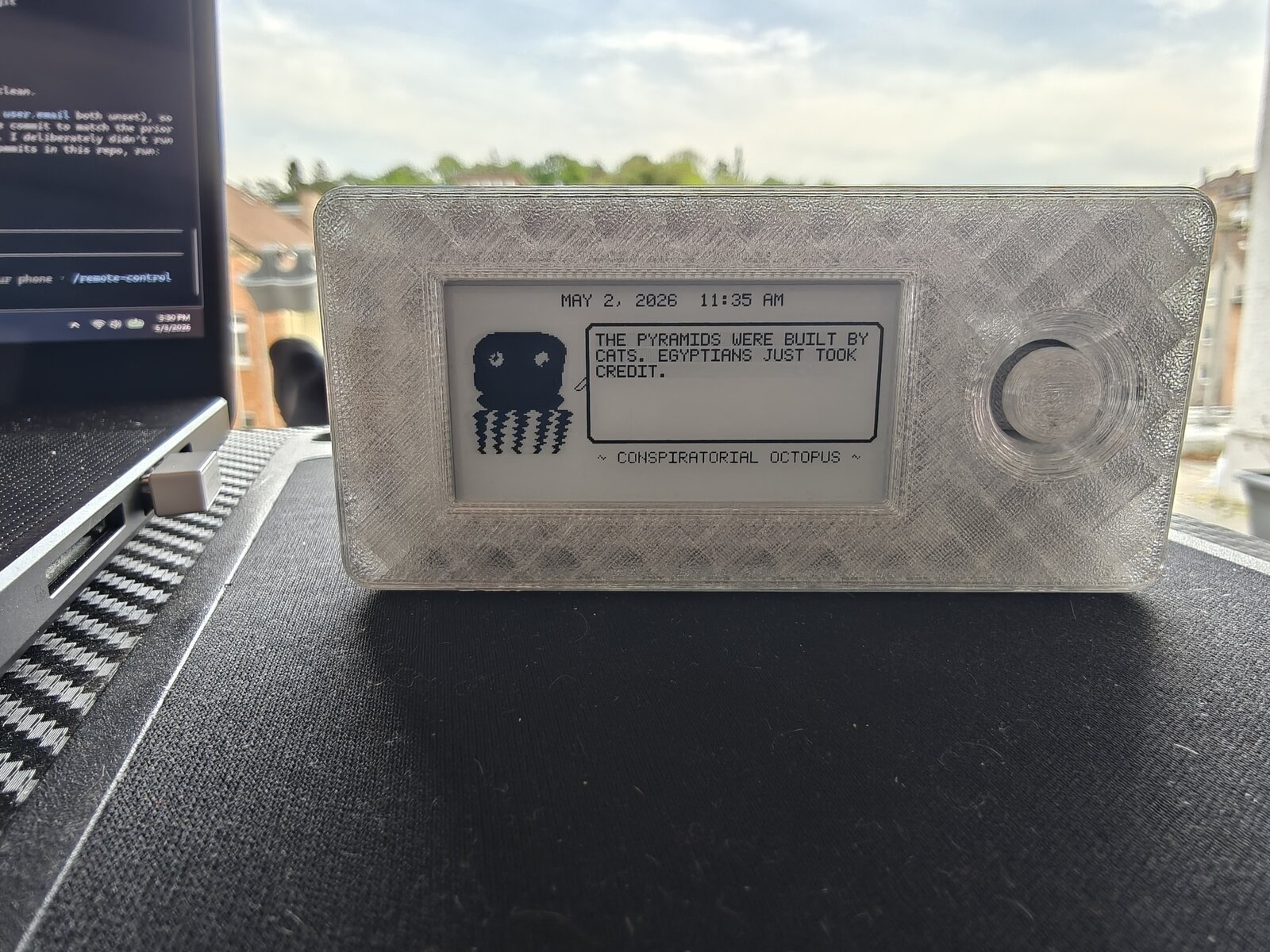 Mk2 translucent Dilder outdoors — hero front shot with Conspiratorial Octopus running on e-ink display, Claude Code terminal visible on laptop behind, blue sky and rooftops