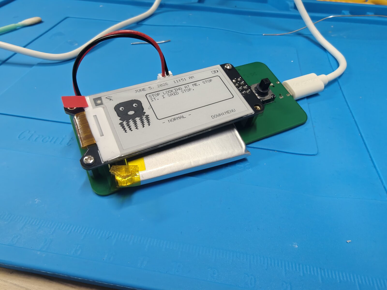 Dilder Board running firmware on the bench — e-ink reads "STOP LOOKING AT ME. STOP IT. I SAID STOP." with the octopus, clock, and NORMAL mood