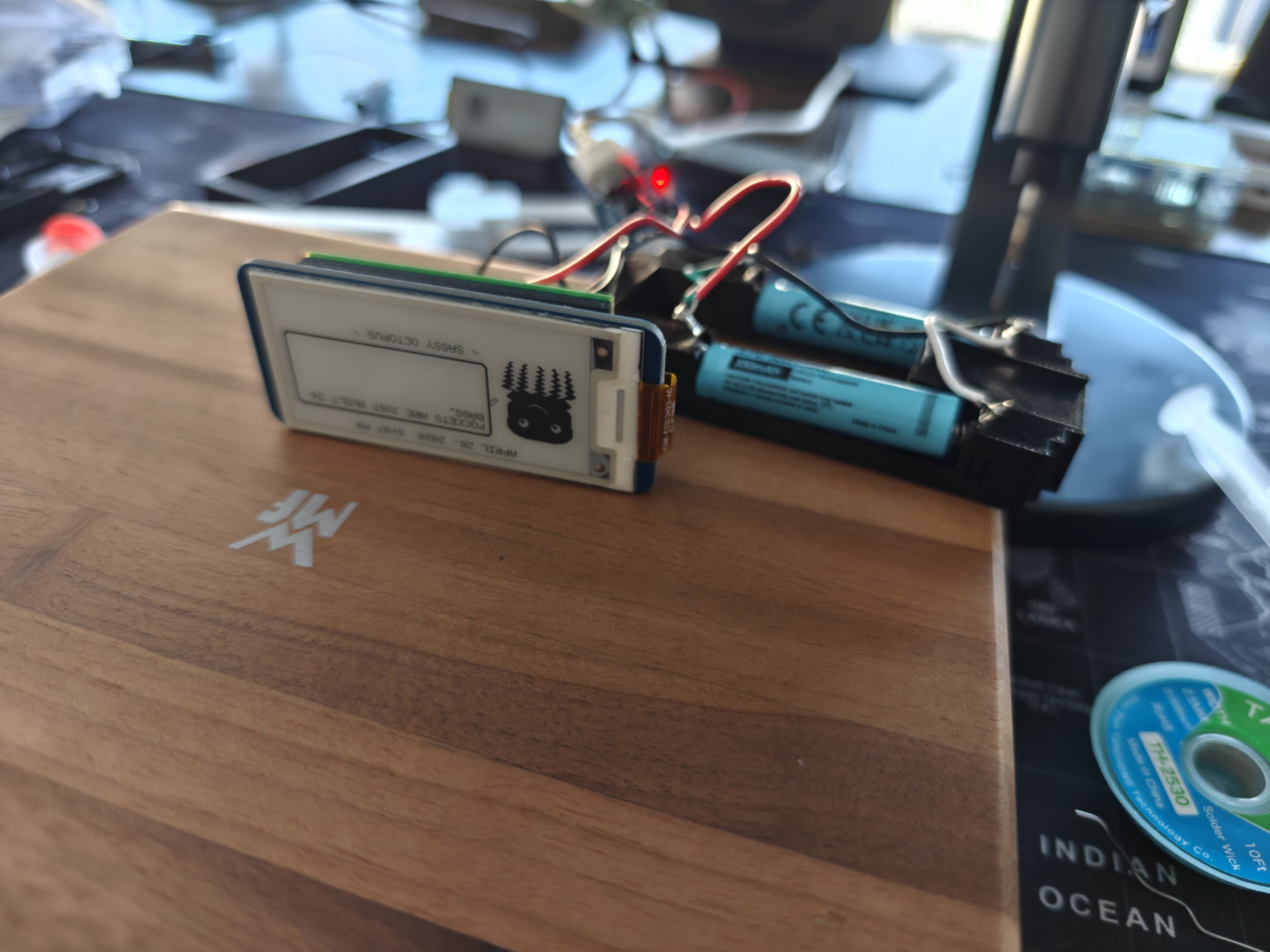 Dilder powered on for the first time on battery — e-ink display showing the Sassy Octopus emotion, 10440 battery in the 3D-printed AAA cradle, TP4056 charge board LED glowing red